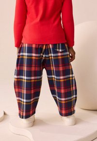 Un top roșu cu mânecă lungă, asortat cu pantaloni largi în carouri navy și roșie, având dungi galbene și albe și un buzunar la spate. Adidașii albi completează ținuta.