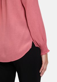 Lys rosa langærmet blouse med struktureret stof, samlede manchetter og sideslids, parret med sorte bukser. Blød, svævende design.