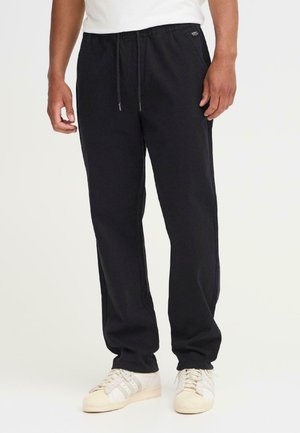 Black cotton jogger pants with an elastic waistband, drawstring, side pockets, and a relaxed fit, paired with white sneakers.