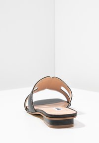 Black leather sandal with an open design, flat heel, and beige inner sole. Features cutout accents and contrasting stitching detail.