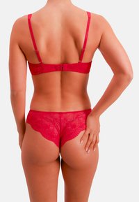 Punainen pitsinen alusasusetti, jossa on rintaliivit säädettävillä olkaimilla ja takana hakaskiinnitys, yhdistettynä vastaaviin läpikuultaviin boyshort-mallisiin alushousuihin, joissa on aaltoilevat reunat.