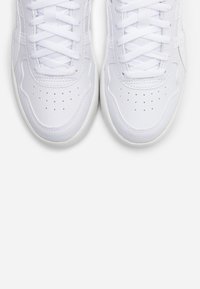 White athletic sneakers with a textured leather upper, rounded toe, flat laces, and perforations for ventilation on a light background.