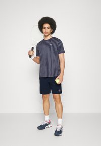 Marine stripete treningsskjorte med logo, kombinert med marineblå shorts. Holder en tennisracket og tennisball, iført sportsko og sokker.