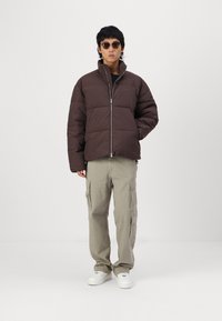 Doudoune marron avec un col haut et une fermeture éclair, associée à un pantalon cargo clair et des baskets blanches. Des lunettes de soleil noires complètent la tenue.
