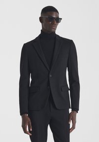 Giacca nera slim fit, con design monopetto, due tasche frontali e bottoni. Indossata sopra un dolcevita nero. Occhiali da sole neri.