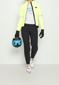 Heldere neon gele fietsjack met reflecterende accenten, zwarte lichtgewicht broek, zwarte handschoenen en een blauw-zwarte helm. Witte schoenen met een patroon.