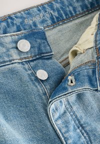 Detail modrých riflí s dvoma zapnutými kovovými gombíkmi a "Flare Fit" vytlačeným vo vnútri pásu.