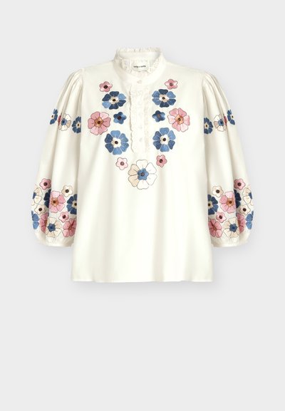 Blouse blanche à manches longues avec col en dentelle et patte de boutonnage, décorée de motifs floraux brodés roses, bleus et crème sur le devant et les manches.