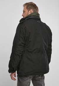 Zwarte jack met een ritskraag, verstelbare tailleband en patches. Vergezeld van een groene sjaal met patroon. Gelaagd materiaal met een matte afwerking.