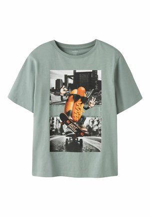 Groen T-shirt met een afbeelding van een hotdog met een zonnebril die aan het skateboarden is met stadsgebouwen op de achtergrond.