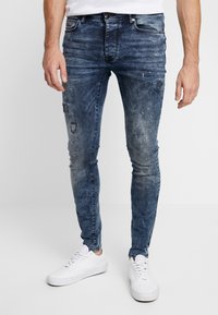 Vaqueros de denim azul de corte ajustado con detalles desgastados, lavado descolorido y cremallera, combinados con zapatillas blancas.