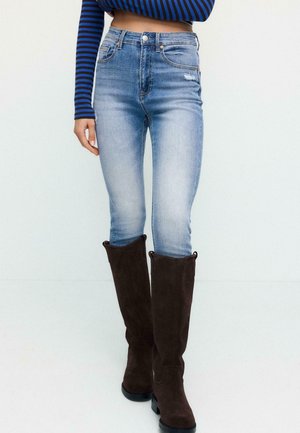 Frau trägt hellblaue Skinny-Jeans, ein bauchfreies langärmliges blau-schwarz gestreiftes Oberteil und kniehohe dunkelbraune Wildlederstiefel.