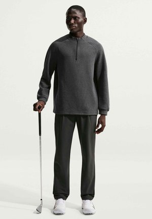 Homme portant un pull à fermeture éclair quart et un pantalon noir, tenant un club de golf, debout devant un fond clair uni.