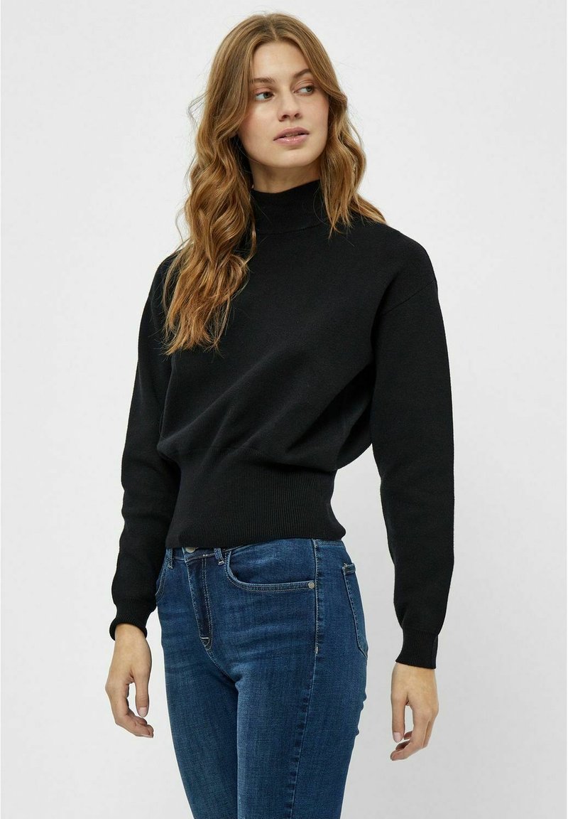 Zwarte gebreide coltrui met lange mouwen en ribbelboord, gecombineerd met blauwe jeans. Zachte textuur, ontspannen pasvorm, minimalistisch ontwerp.
