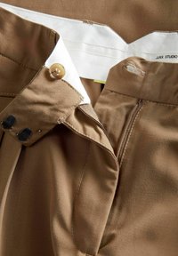 Bruine stoffen broek met een witte tailleband, voorzien van een knoopsluiting, rits en zijzakken. Zichtbare stikseldetails.