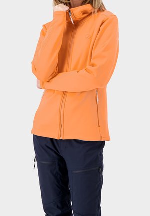 Personne portant une veste orange à fermeture éclair avec un col montant et un pantalon bleu marine, debout les bras croisés et une main touchant le col.