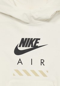 Balta medvilninė džemperis su juodu "NIKE" logotipu ir "AIR" tekstu, žemiau raidžių esantis smėlio spalvos dryžuotas dizainas. Klasikinis pertempiamas stilius.