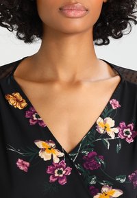 Blouse noire à fleurs avec un col en V, ornée de fleurs violettes, jaunes et orange vibrantes, et un tissu noir transparent sur les épaules.