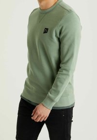 Uomo che indossa un felpa a maniche lunghe verde chiaro con un piccolo logo nero sul petto, abbinata a pantaloni neri, in piedi contro uno sfondo chiaro.