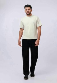 Homem com cabelo e barba escuros, vestindo uma camisa de mangas curtas bege claro, calças pretas e ténis pretos, caminhando para a frente num fundo simples.