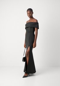 Robe noire à épaules découvertes avec une fente haute au cuisse, corsage ajusté et tissu texturé. Associée à des talons bleu marine et un petit sac à main noir.