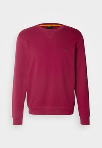 Sweatshirt à manches longues rouge profond avec col rond, poignets et ourlet côtelés, et petit écusson logo carré sur le côté gauche de la poitrine.