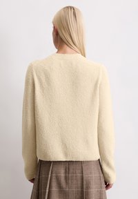 Kremowy, teksturowany sweter z okrągłym dekoltem i długimi rękawami, w połączeniu z brązową spódnicą w kratę. Skupienie na wykonaniu i dopasowaniu.