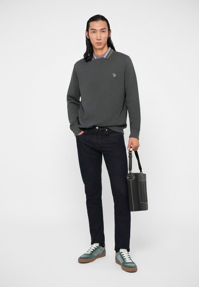 Vyras, apsirengęs tamsiai žaliu megztiniu virš striukinio marškinių, tamsiomis džinsinėmis kelnių ir cyano spalvos sportbačiais, laiko juodą krepšį su dirželiu prieš baltą foną.