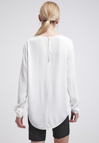 Vit långärmad blus med rundade kanter, sprund på baksidan och slät textur. Ärmarna har manschetter med knappar. Buren tillsammans med svarta shorts.