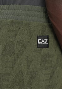 Tuvumā attēloti olīvzaļi flīsa bikses ar EA7 Emporio Armani logotipa uzplecu uz augšstilba un elastīgu jostasvietu.