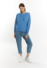 Felpa blu chiaro con cappuccio regolabile, vestibilità over size, polsini a costine, abbinata a jeans chiari con risvolto e sneaker bicolore.