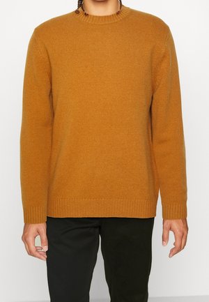Persoon in een mosterdkleurige gebreide trui met lange mouwen en zwarte broek, staand tegen een effen lichte achtergrond.