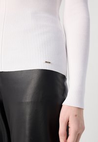 Haut long à manches blanches côtelé associé à un pantalon en cuir noir lisse, avec un petit accent de logo métallique près de l'ourlet.
