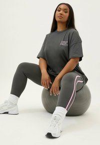 Grå oversized t-shirt med tryckt text, grå leggings med rosa sidosträck, vita träningsskor, sittande på en grå träningsboll.