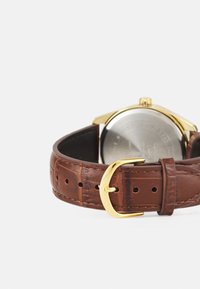 Brown leather strap with crocodile texture, gold-tone buckle, and a round case visible from the back, revealing the silver watch face.