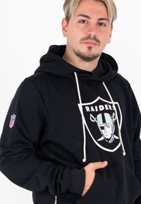 Zwarte hoodie met een voorzak, voorzien van een Raiders-logo in wit en zilver. Witte koordjes en een NFL-patch op de mouw. Zacht stof.