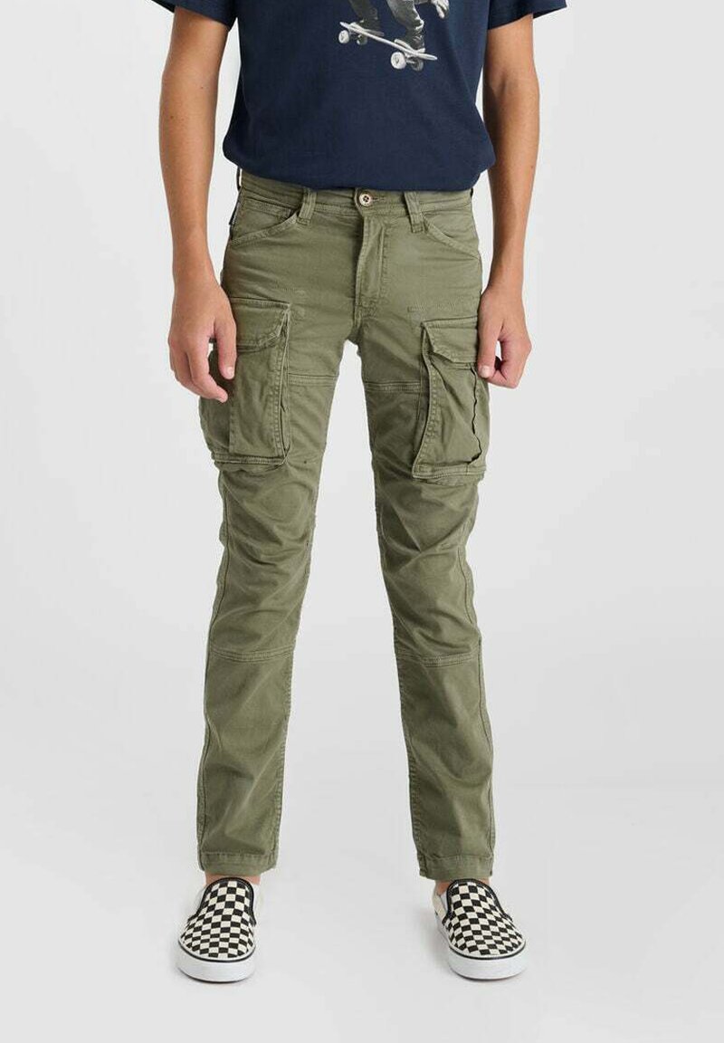Personne portant un pantalon cargo vert olive, un t-shirt graphique de skateboard bleu marine, et des chaussures slip-on à damiers noir et blanc, debout devant un fond uni.