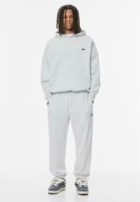 Felpa grigia con cappuccio e pantaloni della tuta in un materiale morbido, con un logo nero sul petto e sulla gamba. Il modello indossa sneakers grigie e blu.
