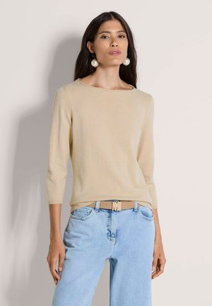 Vrouw die een beige top met lange mouwen draagt, lichtblauwe spijkerbroek, beige riem met een "M" gesp en grote ronde pareloorbellen, staand tegen een effen lichte muur.