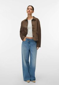 Veste courte marron avec fermeture à boutons, deux poches avant, associée à un jean large bleu clair. Le mannequin porte un haut blanc ajusté.