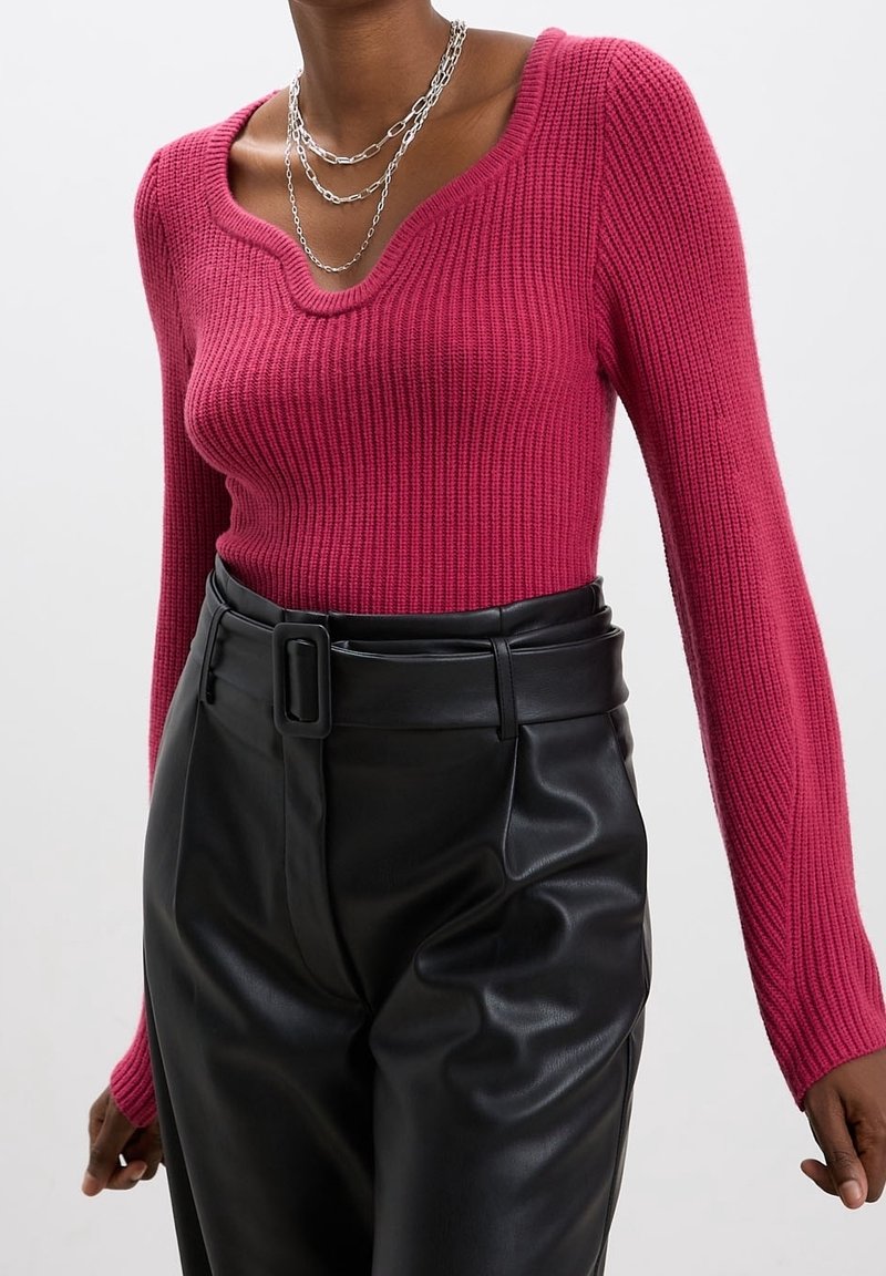 Femme portant un pull rose côtelé à manches longues, un pantalon noir taille haute en simili cuir avec une ceinture, et des colliers en chaîne argentée superposés.