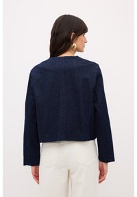 Chaqueta de mezclilla azul marino, diseño corto, mangas largas, textura suave, con un cuello simple. Combinada con pantalones blanco roto en el fondo.