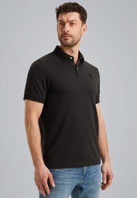 Zwarte polo met een kraag, korte mouwen en een subtiel logo. Gemaakt van een soepele stof, voorzien van een knoopsluiting. Draagt met blauwe jeans.
