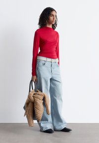 Rotes langärmliges Oberteil, hellblaue weit geschnittene Jeans, weißer Gürtel, schwarze Mules, hält einen hellbraunen Strickpullover und eine schwarze Tote-Tasche.