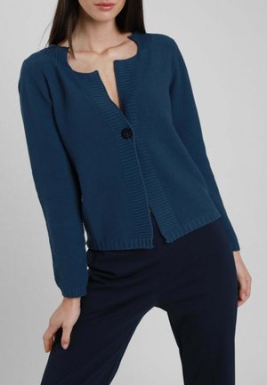 Donna che indossa un cardigan blu scuro a trama con un solo bottone nero allacciato, abbinato a pantaloni blu scuro coordinati su uno sfondo neutro.