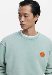 Sweatshirt vert menthe clair en tissu doux avec un col côtelé et un logo de fleur orange sur la poitrine gauche. Coupe décontractée avec des épaules tombantes.