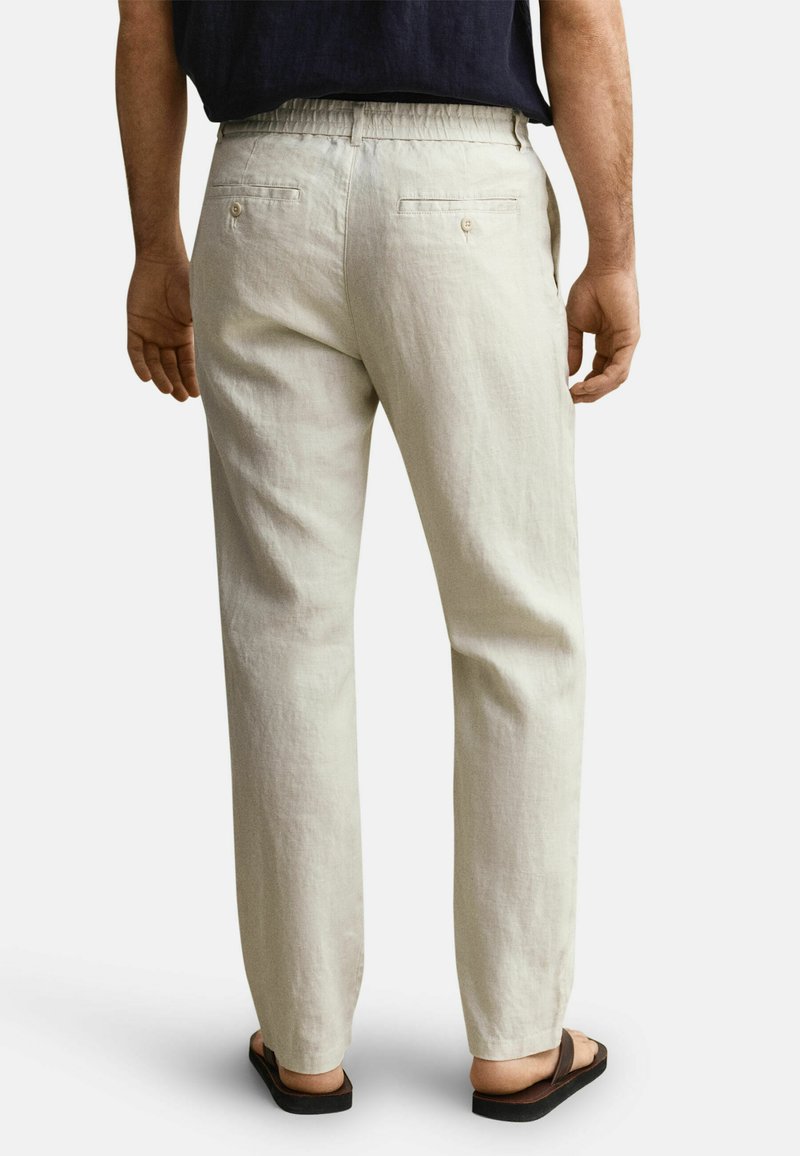 Man draagt lichtbeige casual broek met achterzakken en donkere sandalen, staand tegen een effen lichte achtergrond.