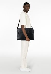 Sort læder messenger taske med en struktureret design, top lynlås lukning, forlommes og justerbar rem, holdt af en person i et lyst outfit.