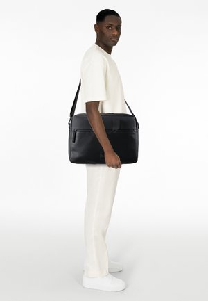 Schwarze Leder-Umhängetasche mit strukturiertem Design, oberem Reißverschluss, Fronttasche und verstellbarem Riemen, gehalten von einer Person in einem hellen Outfit.