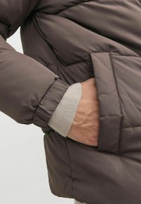 Giacca puffer marrone con una finitura opaca e testurizzata. Presenta polsini a costine e tasche laterali. I dettagli cuciti aggiungono struttura.